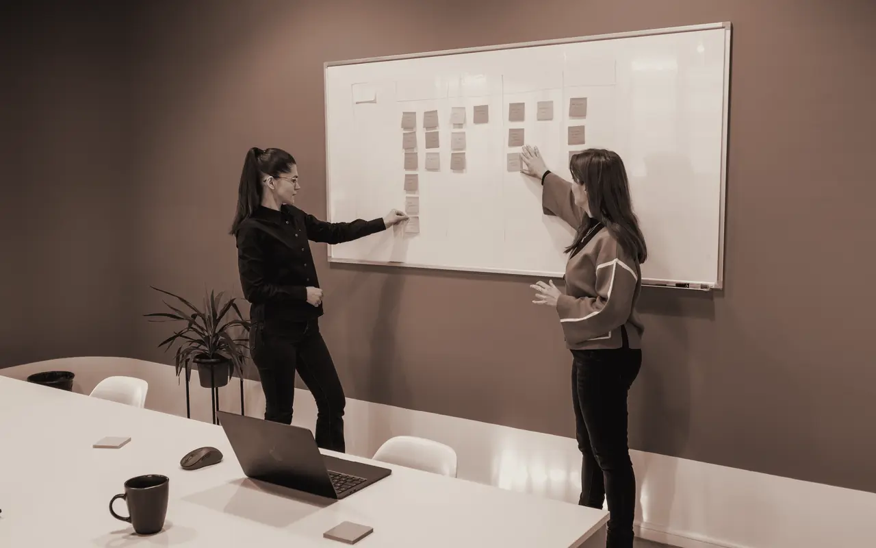 Dos personas trabajan de pie frente a una pizarra blanca con notas adhesivas.